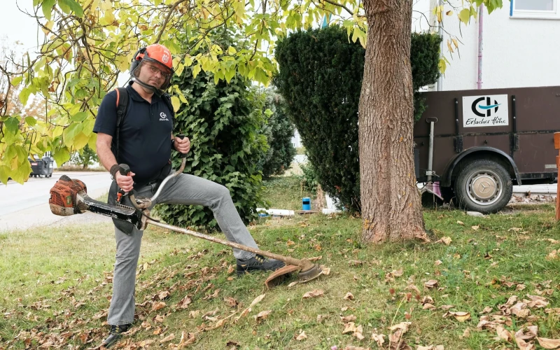 Mann mit Motorsense steht auf einem grasbewachsenen Hang neben einem Baum und schneidet Gras. Im Hintergrund ist ein Anh&auml;nger mit dem Logo der Erlacher H&ouml;he zu sehen.