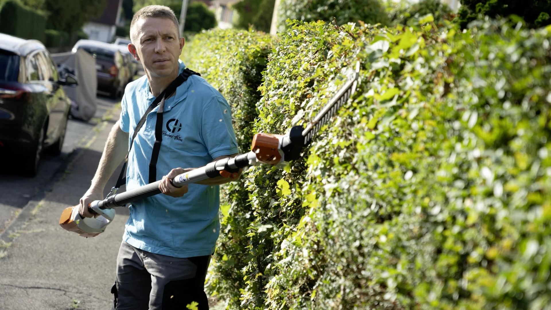 Ein Mann im blauen T-Shirt mit Logo der Erlacher H&ouml;he steht auf einem Gehweg und schneidet eine gr&uuml;ne Hecke mit einer Heckenschere. Im Hintergrund sind parkende Autos und H&auml;user zu sehen sind.