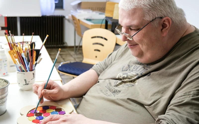 Ein Mann mit Brille sitzt an einem Tisch und h&auml;lt einen Pinsel in der Hand mit dem er bunte Pl&auml;ttchen in einer Holzblumen anordnet. Neben ihm stehen mehrere Pinsel in einem Becher und im Hintergrund ein weiterer Tisch mit St&uuml;hlen. Er tr&auml;gt ein graues T-Shirt und hat Freude an seiner kreativen Besch&auml;ftigung.