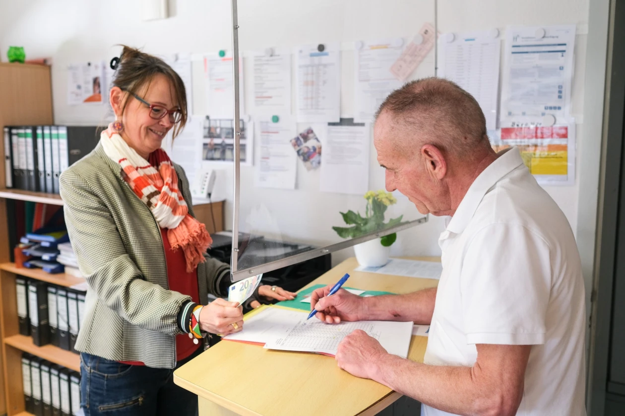 Eine Frau mit orange-wei&szlig;en Schal und grauem Blazer steht an einem Schreibtisch und h&auml;lt 20 Euro in der rechten Hand. Ein Mann mit wei&szlig;em T-Shirt steht auf der anderen Tischseite mit einem Kugelschreiber in der Hand und unterschreibt ein Dokument auf den Tisch. Im Hintergrund stehen Ordner in Regalen und an der Wand h&auml;ngen Dokumente.