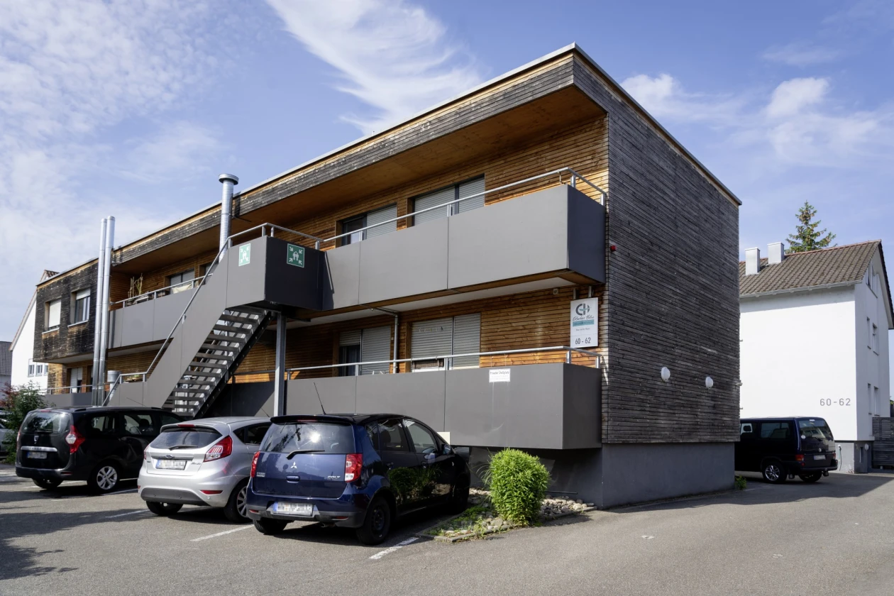Modernes Zweistockgeb&auml;ude mit grauer Fassade und Holzbeplankung, ausgestattet mit Au&szlig;entreppe und Parkpl&auml;tzen mit parkenden Autos. Die Architektur verbindet zeitgen&ouml;ssisches Design mit funktionaler Gestaltung und bietet einen klaren, strukturierten Eindruck.