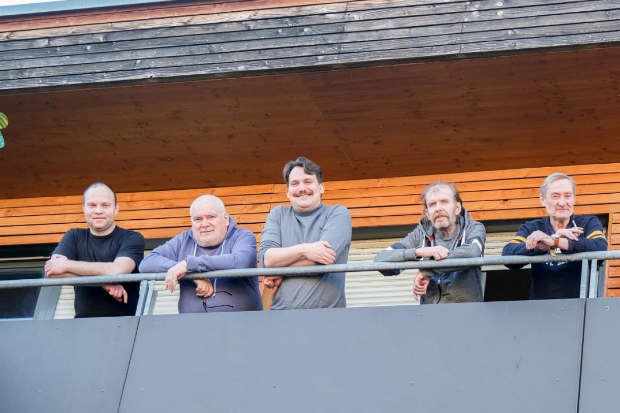 Sechs M&auml;nner und eine Frau stehen auf einem Balkon, lehnen sich ans Gel&auml;nder und blicken l&auml;chelnd in die Kamera.