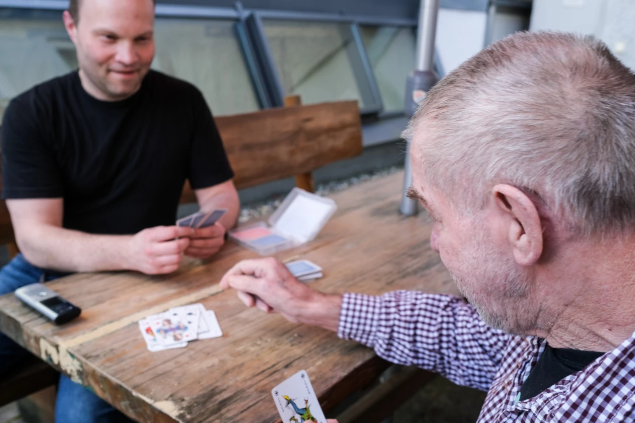 Zwei M&auml;nner sitzen mit ausgelegten Spielkarten an einem Holztisch. Der Mann in kariertem Hemd sitzt mit dem R&uuml;cken zur Kamera, der andere Mann, der ihm gegen&uuml;ber sitzt tr&auml;gt ein schwarzes T-Shirt.