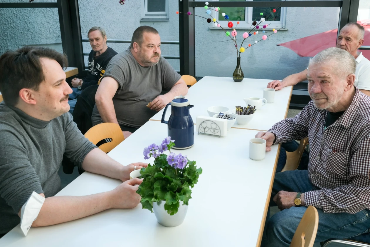 Mehrere Personen sitzen kaffeetrinkend an einer Tischreihe mit einer gr&uuml;nen Pflanze. Im Hintergrund ist eine Fensterfront zu sehen.