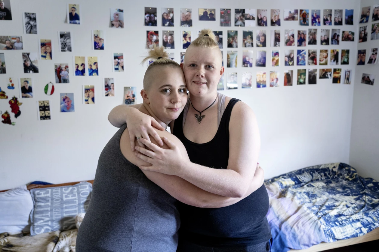 Zwei Frauen umarmen sich herzlich in einem Zimmer. Im Hintergrund steht ein Bett an der Wand, die mit vielen Fotos geschm&uuml;ckt ist. Beide tragen dunkle Tops und schauen direkt in die Kamera.