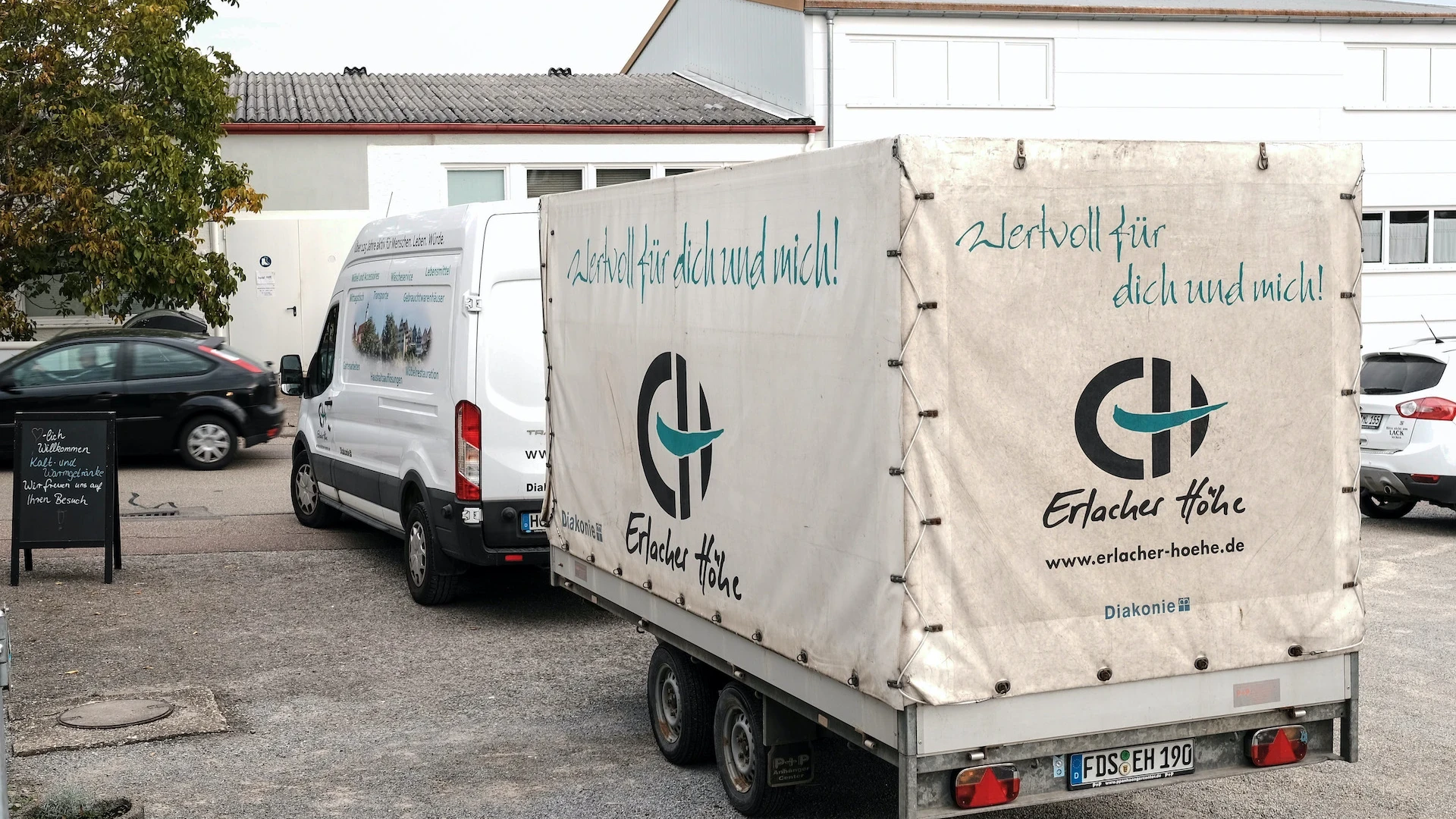 Ein wei&szlig;er Transporter mit Anh&auml;nger steht vor einem Geb&auml;ude auf dem Hof. Auf dem Transporter ist das Logo der Erlacher H&ouml;he zu sehen.