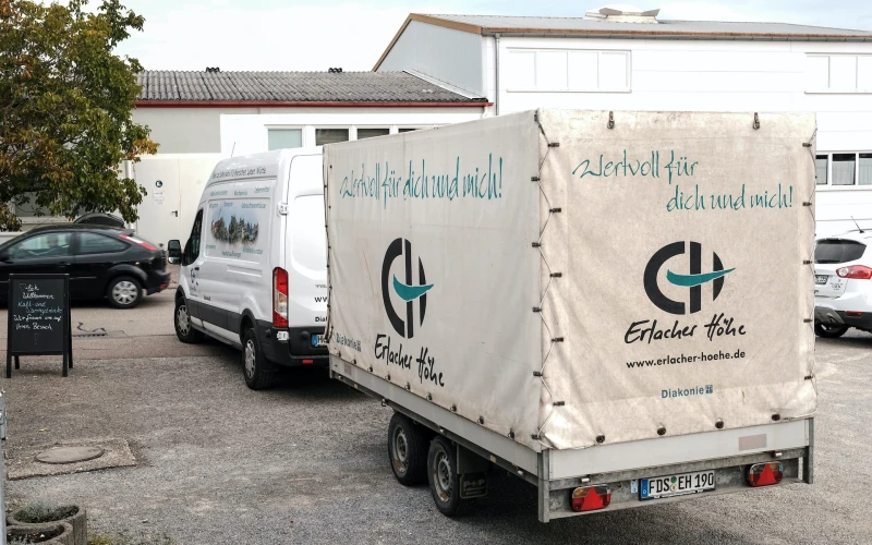 Ein wei&szlig;er Transporter mit Anh&auml;nger steht vor einem Geb&auml;ude auf dem Hof. Auf dem Transporter ist das Logo der Erlacher H&ouml;he zu sehen.