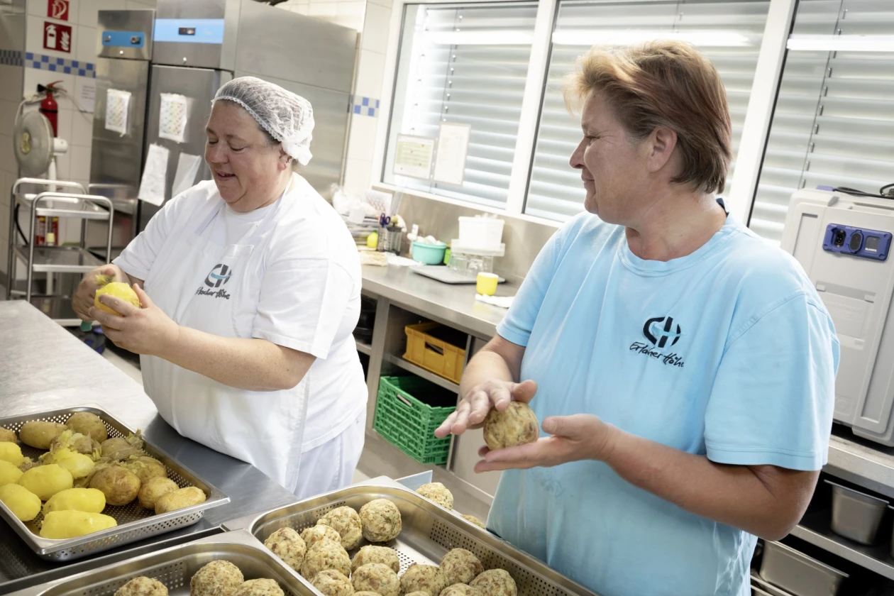 Zwei Frauen stehen in einer K&uuml;che. Eine Frau tr&auml;gt ein wei&szlig;es T-Shirt, die Frau ein hellblaues T-Shirt, beide mit Logo der Erlacher H&ouml;he. Die linke Frau sch&auml;lt Kartoffeln, die rechte Frau formt Semmelkn&ouml;del. Sie stehen an einem Arbeitstisch mit Metallbeh&auml;ltern.