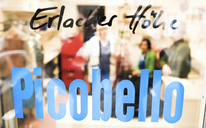Glast&uuml;re mit dem blauen Schriftzug "Picobello" und dem Text "Erlacher H&ouml;he" in schwarzer Schrift. Beim Durchblicken sind verschwommen Menschen zu sehen.