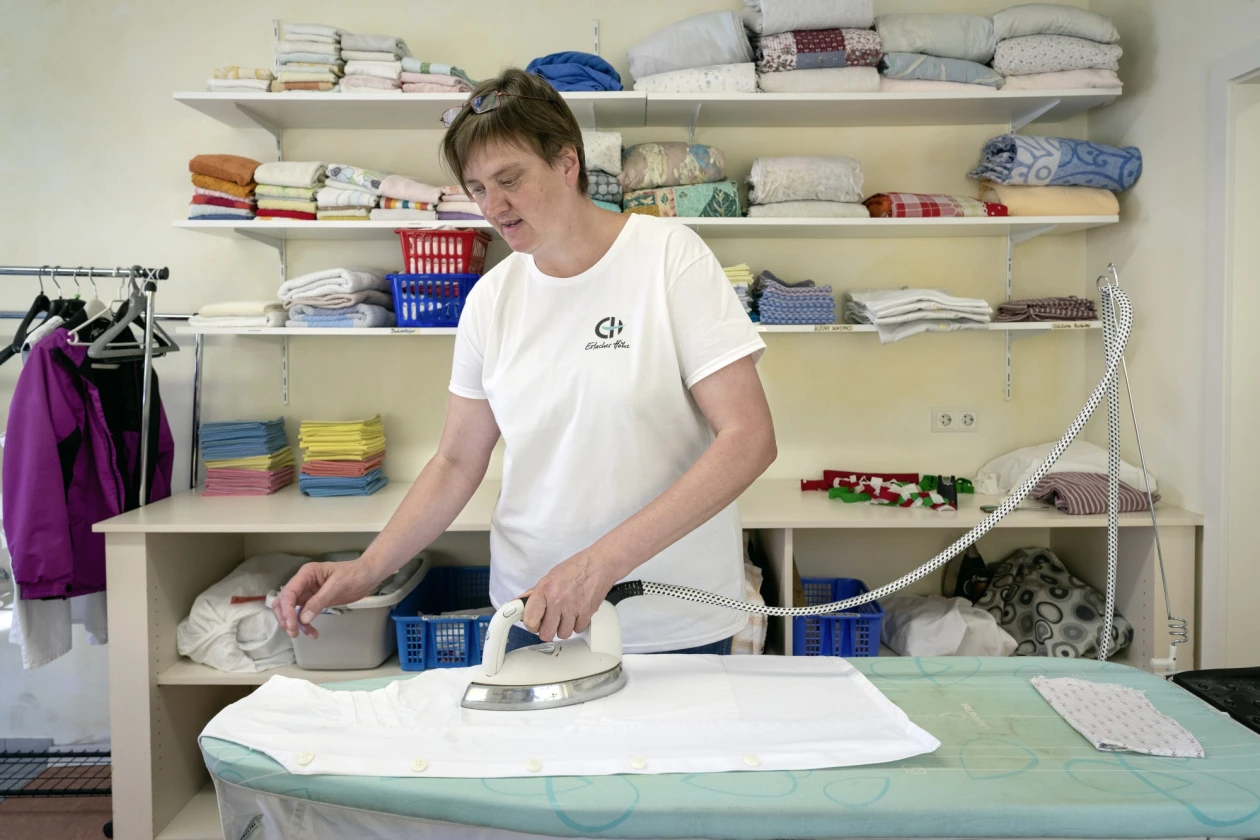 Eine Frau in wei&szlig;em T-Shirt und Erlacher H&ouml;he Logo beim B&uuml;geln eines wei&szlig;en W&auml;schest&uuml;cks. Im Hintergrund ist ein Regal mit verschiedenfarbigen Textilien und K&ouml;rben zu sehen.