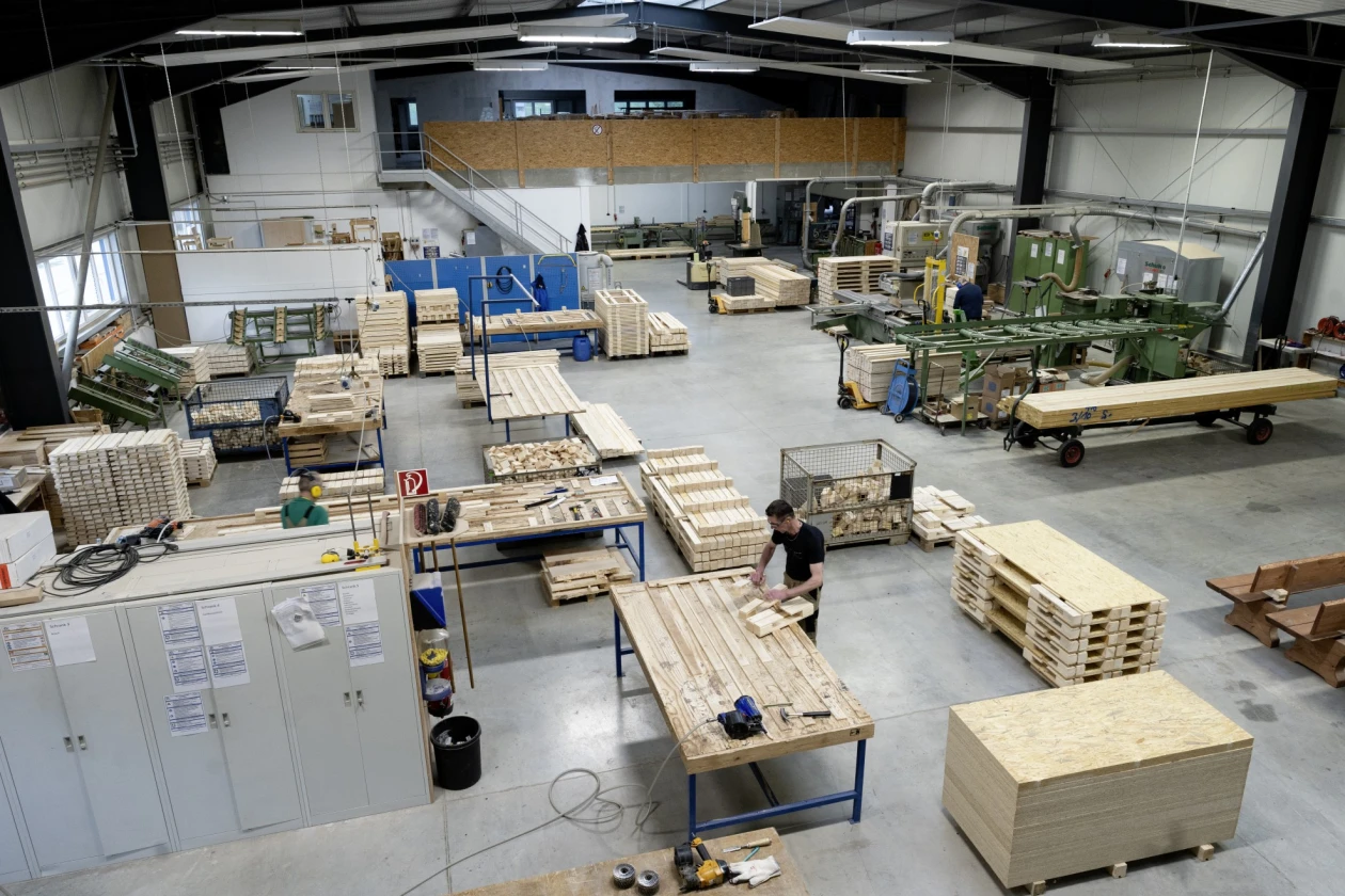 Gro&szlig;e Holzwerkstatt mit verschiedenen Maschinen, Werkb&auml;nken und gestapelten Holzpaletten. Ein Mann steht an einer Werkbank und arbeitet an einem Holzst&uuml;ck. Die Halle hat hohe Decken und ist mit industrieller Ausr&uuml;stung f&uuml;r Holzverarbeitung ausgestattet.