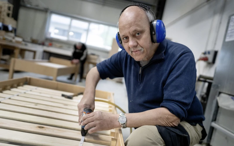 Ein Mann mit blauem Geh&ouml;rschutz l&auml;chelt in die Kamera. Er arbeitet in einer Werkstatt an einem Bettgestell. Im Hintergrund ist eine weitere Person zu sehen, die ebenfalls an einem Bettgestell arbeitet.