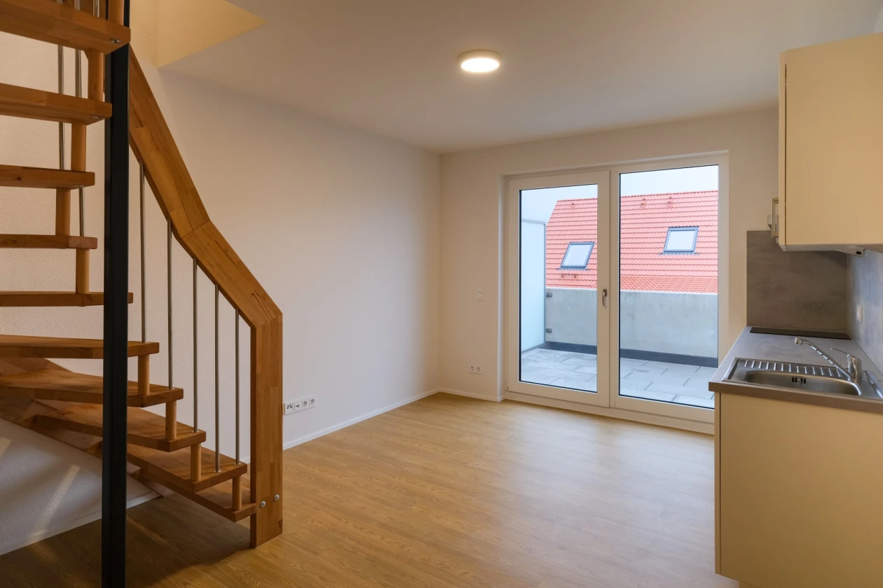 Ein leerer Raum mit Holzboden und einer Holztreppe. Es gibt einen gro&szlig;en Balkon mit einer Fensterfront, und eine kleine K&uuml;chenzeile ist ebenfalls im Raum vorhanden.