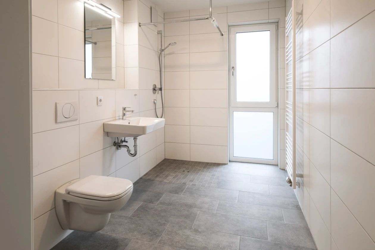 Modernes, helles Badezimmer mit wei&szlig;en Wandfliesen, grauem Steinboden, wandh&auml;ngendem WC und Waschbecken. Gro&szlig;es Fenster und Spiegel sorgen f&uuml;r nat&uuml;rliche Helligkeit und einen offenen, luftigen Eindruck.