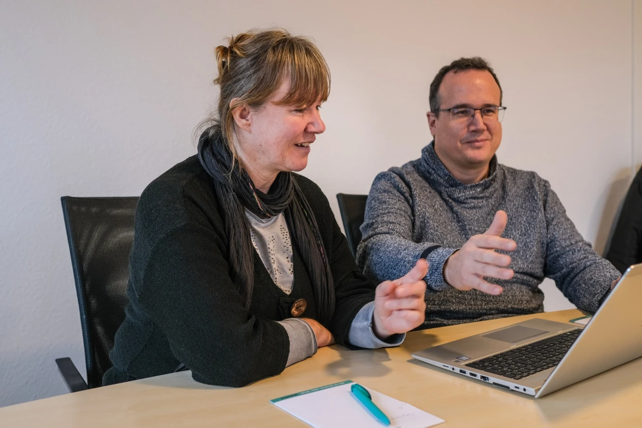 Zwei Personen sitzen an einem Tisch mit einem Laptop: eine Frau in schwarzer Weste mit grauem Unterteil und ein Mann mit grauem Pullover und Brille. Beide l&auml;cheln und schauen auf den Laptop.