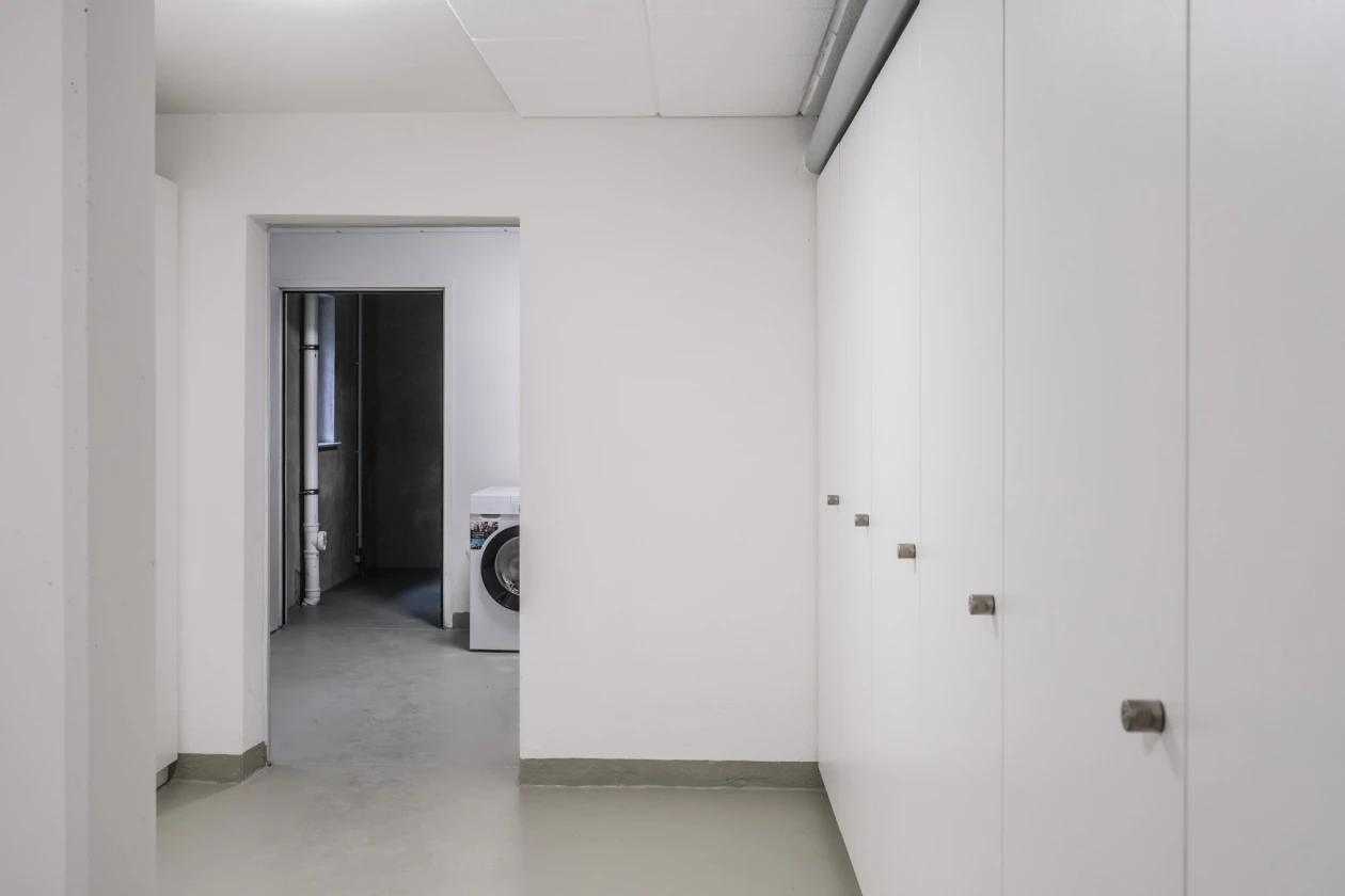 Minimalistischer, wei&szlig;er Flurbereich mit Durchgang zu einem Nebenraum, in dem eine Waschmaschine steht. Glatte W&auml;nde, graue Bodenfliesen und wei&szlig;e Einbauschr&auml;nke verleihen dem Raum eine schlichte, moderne Atmosph&auml;re.