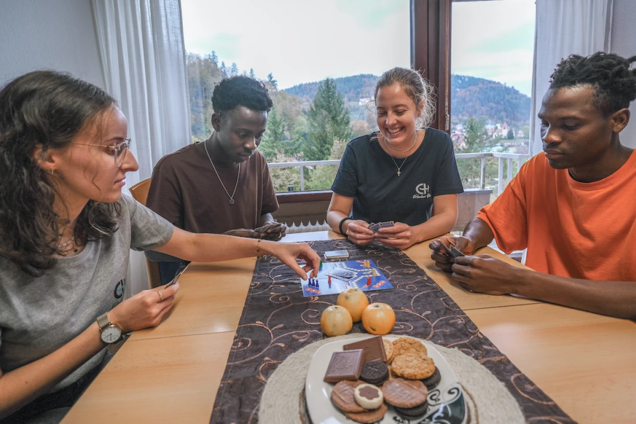 Zwei Frauen und zwei M&auml;nner sitzen an einem Tisch und spielen Mensch &auml;rgere dich nicht, w&auml;hrend im Hintergrund durch ein Fenster Berge und B&auml;ume zu sehen sind. Auf dem Tisch liegt Obst und Geb&auml;ck..