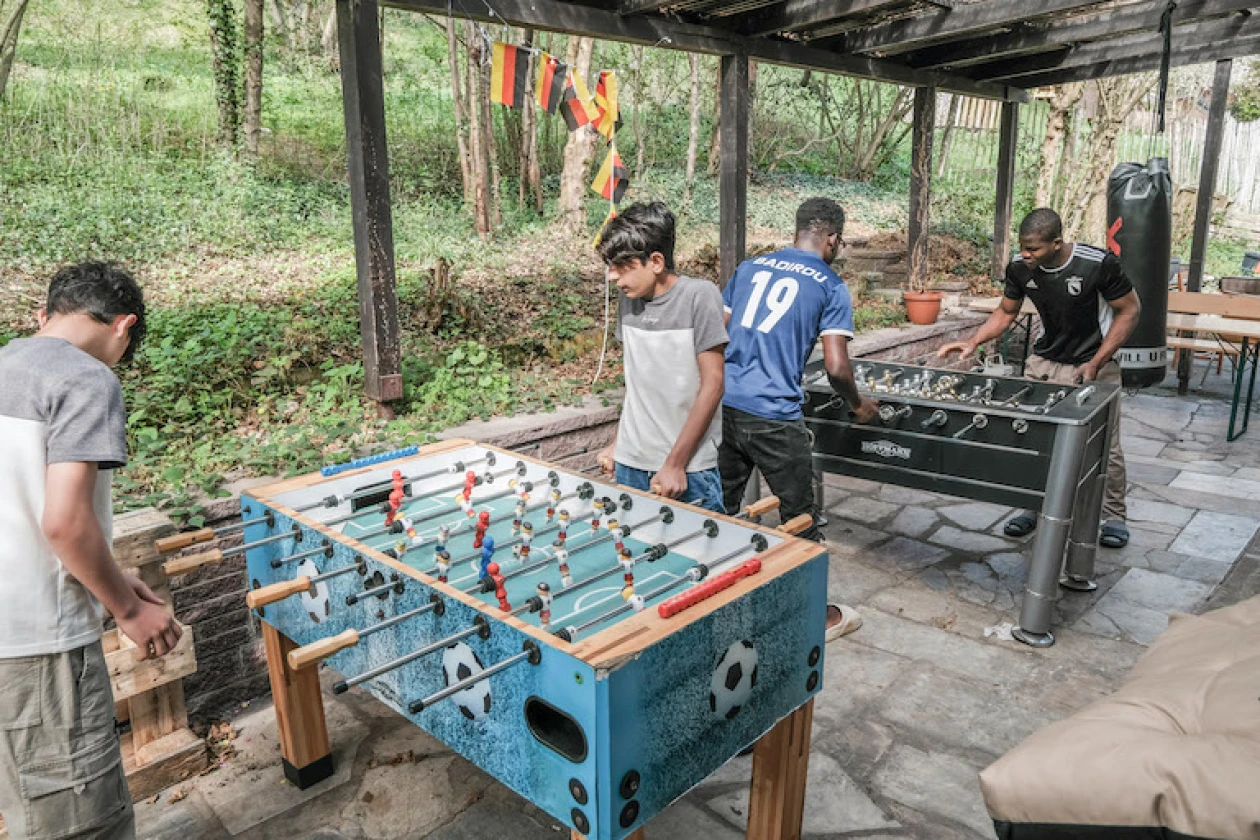 Vier Jugendliche spielen an einem &uuml;berdachten Au&szlig;enbereich Tischkicker. Im Hintergrund sind Holzb&auml;nke und ein Sandsack zu sehen, umgeben von einer gr&uuml;nen Waldkulisse.