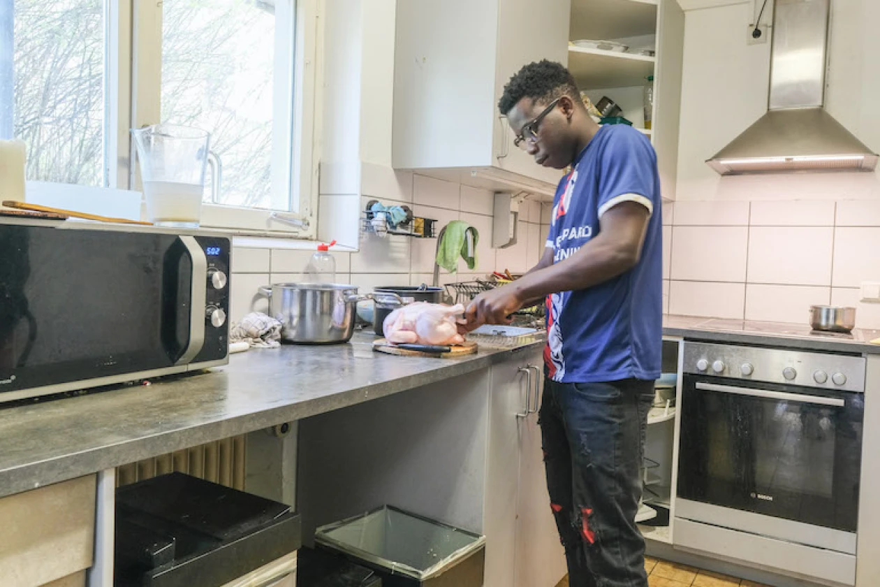 Ein Jugendlicher in blauem T-Shirt bereitet Essen an der K&uuml;chenarbeitsplatte zu, mit Mikrowelle, Backofen und Sp&uuml;le im Hintergrund.