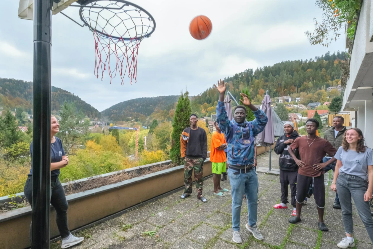 Zwei Frauen und sechs m&auml;nnliche Jugendliche stehen auf einer Terrasse. Ein Jugendlicher in einem blauen Pullover wirft gerade einen Basketball in einen Korb, w&auml;hrend die anderen zuschauen. Im Hintergrund sind H&auml;user und B&auml;ume zu sehen.