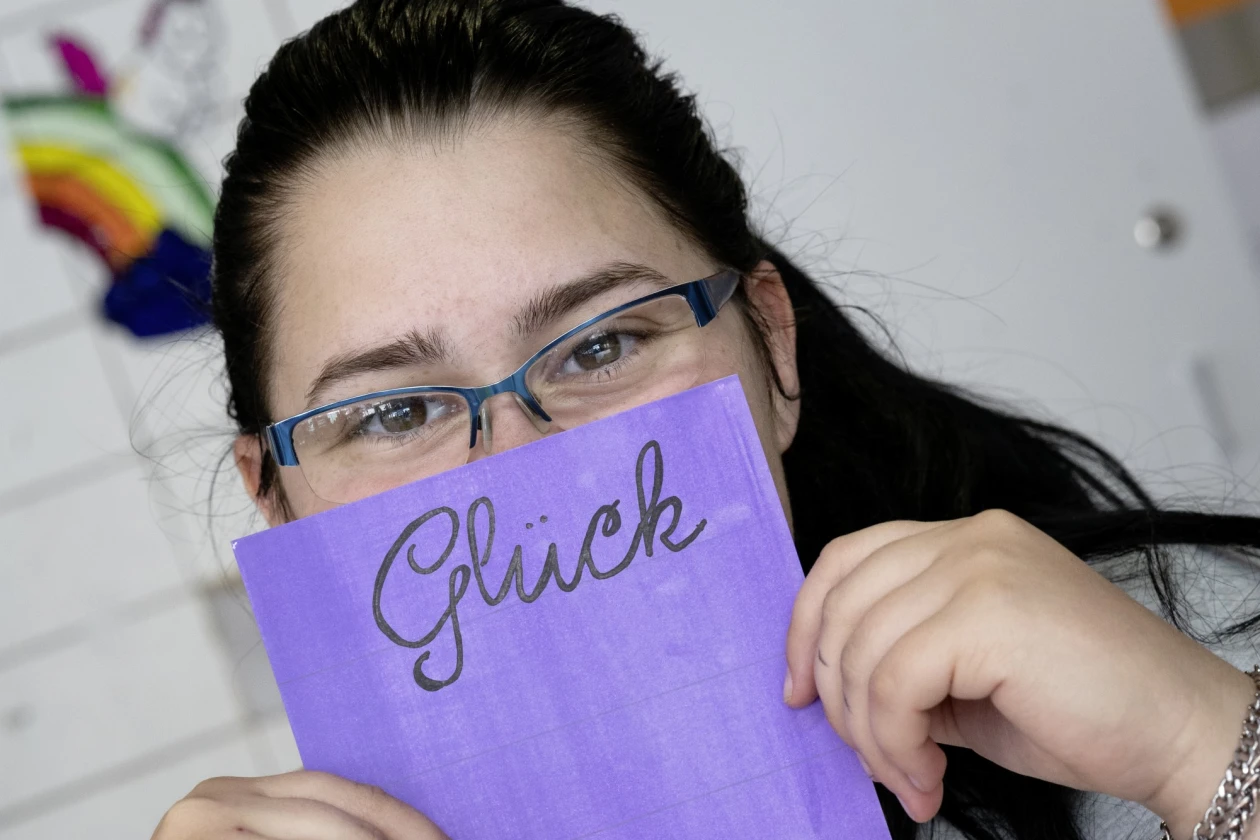 Eine Frau mit Brille tr&auml;gt ein graues T-Shirt mit Aufdruck und h&auml;lt ein violettes Blatt mit dem Wort "Gl&uuml;ck" vor ihren Mund, w&auml;hrend sie an einem Tisch sitzt.