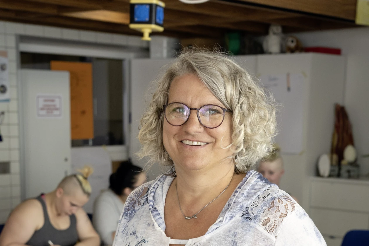 Im Vordergrund steht eine freundlich l&auml;chelnde Frau mit lockigen grauen Haar und Brille. Im Hintergrund sieht man weitere Personen, die an einem Tisch sitzen.