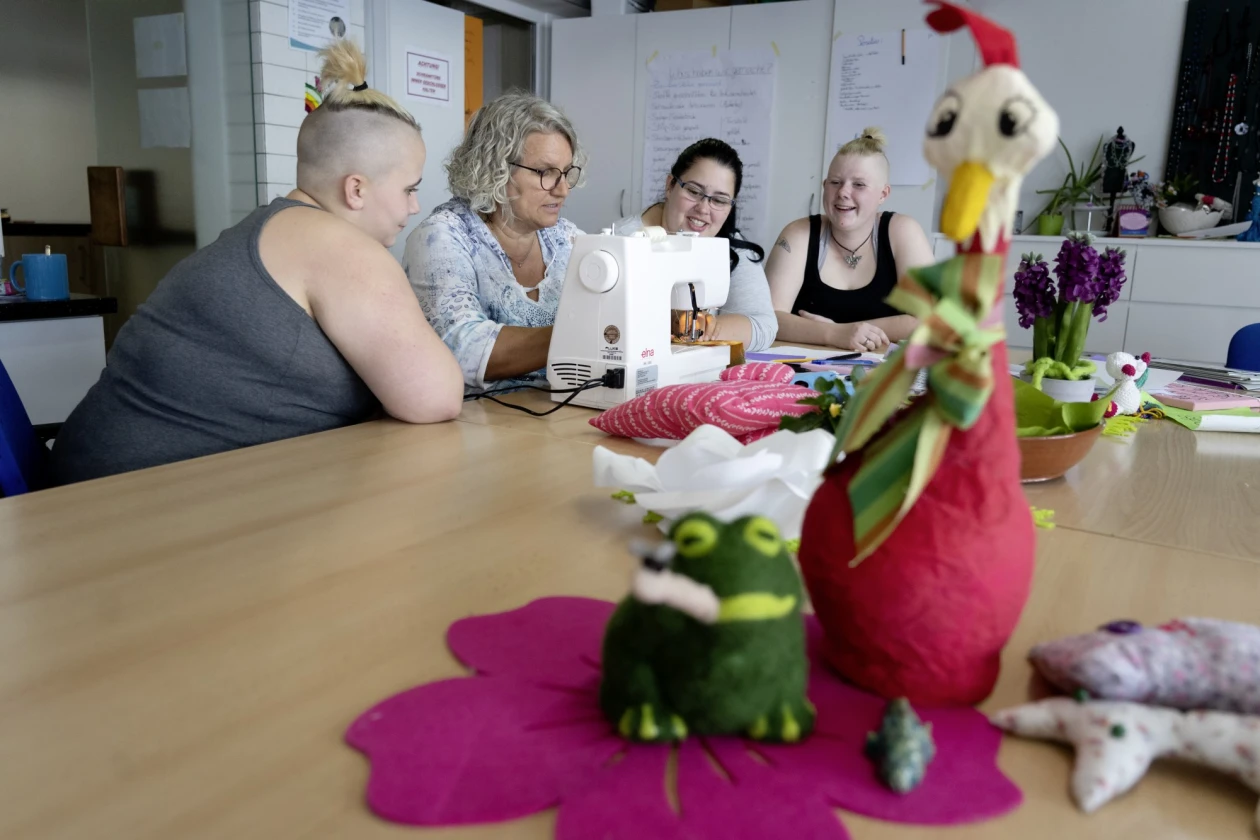 Vier Frauen sitzen an einem Tisch und arbeiten mit einer N&auml;hmaschine, w&auml;hrend im Vordergrund eine rote H&uuml;hnerfigur aus Filz und andere Dekorationsgegenst&auml;nde platziert sind.