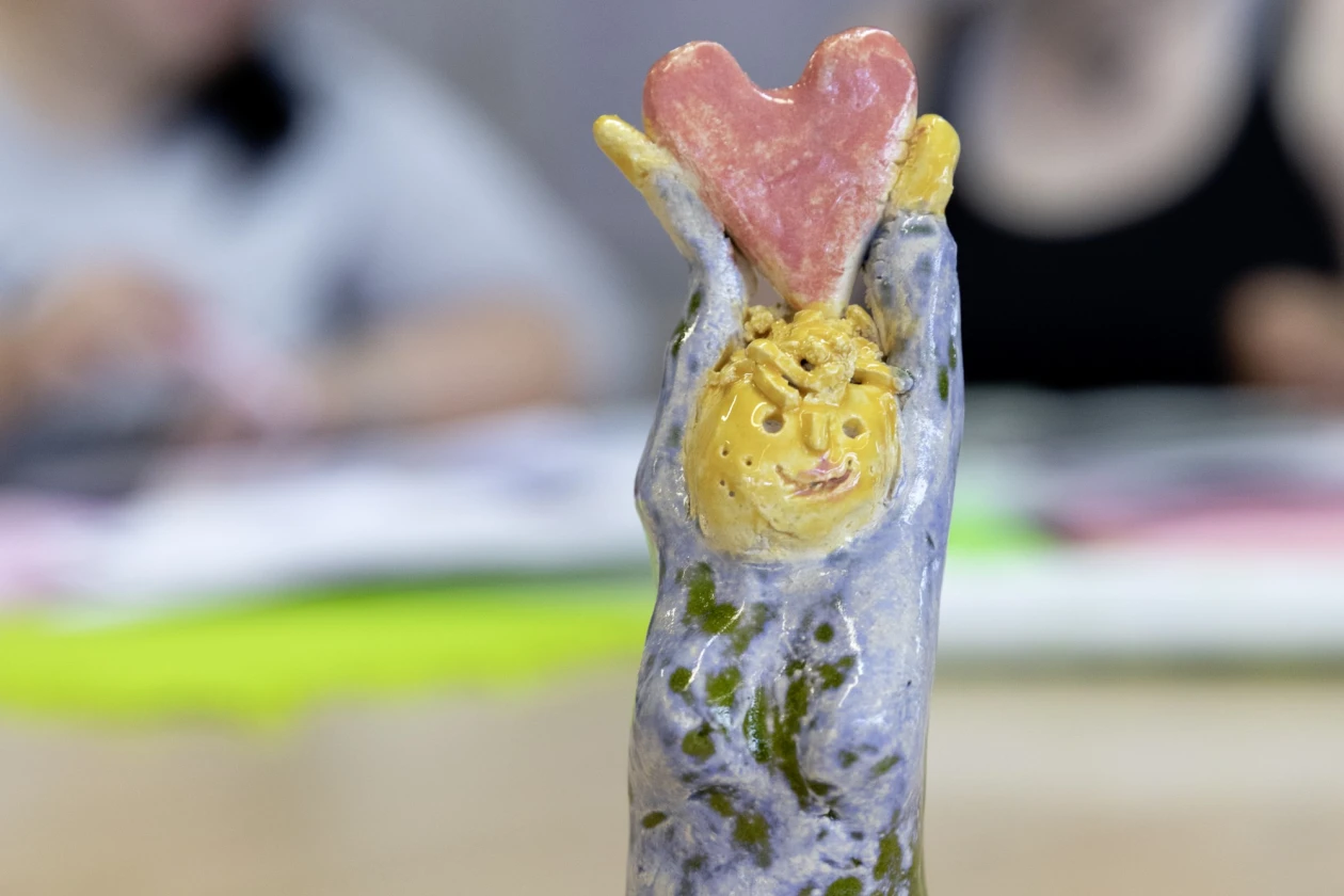Eine handgefertigte Keramikfigur mit gelbem Gesicht und blau-gr&uuml;n meliertem Gewand h&auml;lt ein rotes Herz &uuml;ber dem Kopf. Die Figur steht auf einem Holztisch, im Hintergrund sitzen zwei verschwommene Personen.