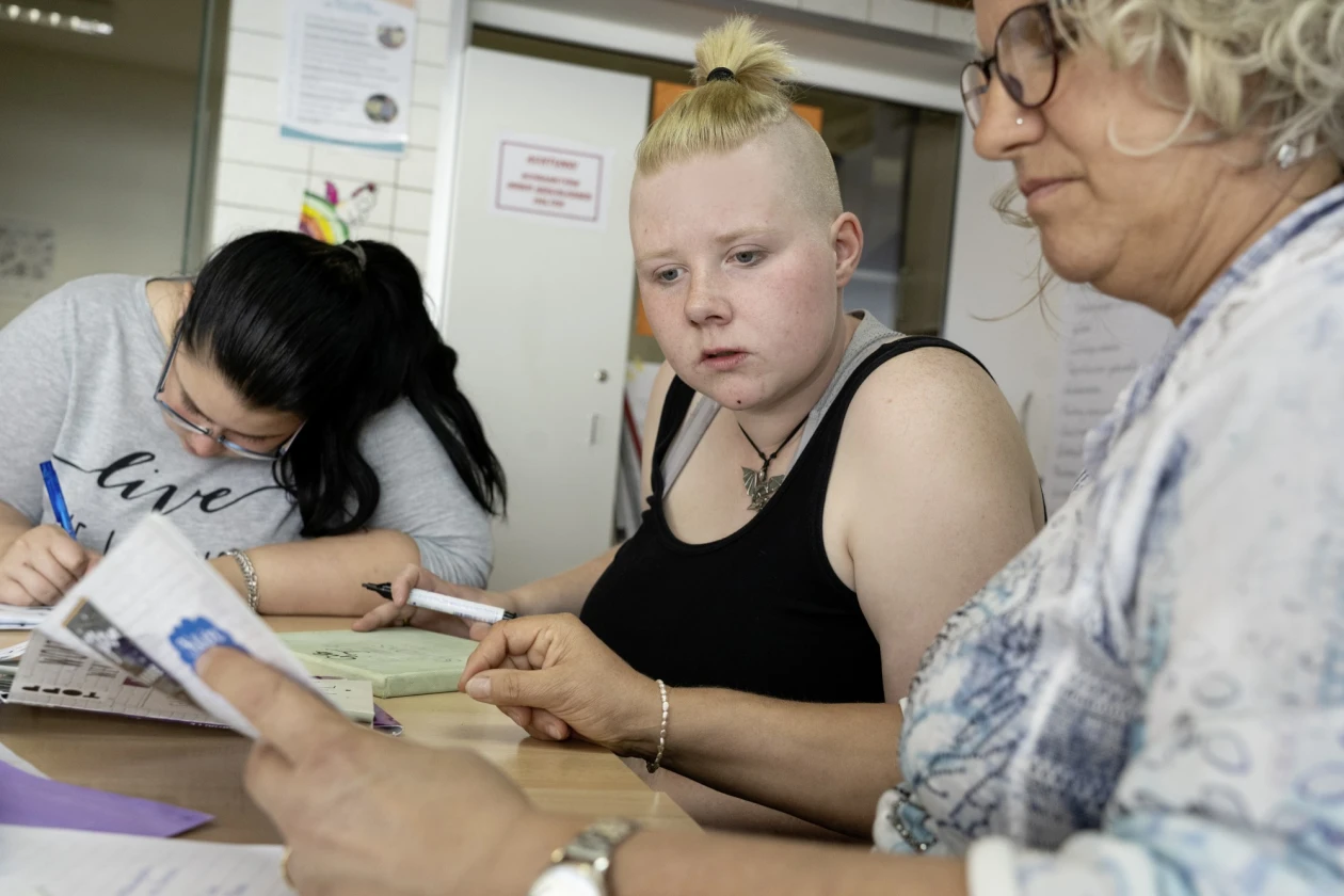 Drei Frauen sitzen an einem Tisch. Eine Frau mit dunklen Haaren schreibt konzentriert, w&auml;hrend die beiden Frauen mit blonden Haaren verschiedene Buntpapiere betrachten.