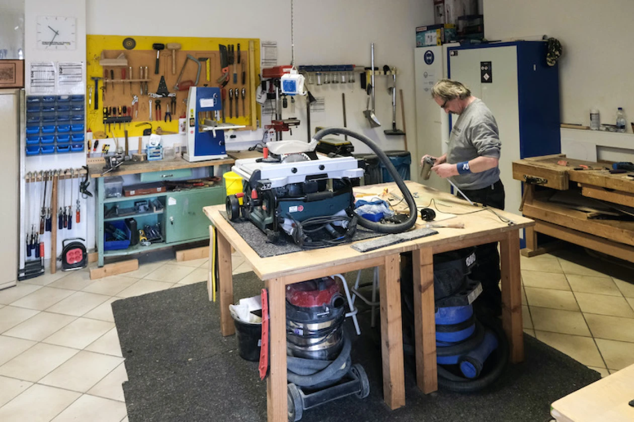 Zu sehen ist ein Raum mit Arbeitsbank, gelber Werkzeugwand und verschiedenen Maschinen. Ein Mann steht an der Werkbank und blickt pr&uuml;fend auf zwei kleine Gegenst&auml;nde, die er in seinen H&auml;nden h&auml;lt.