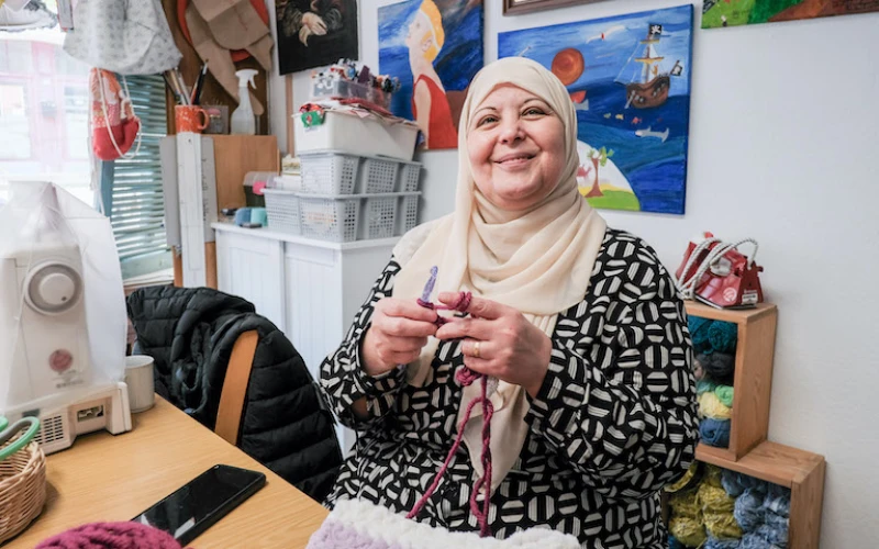 Eine l&auml;chelnde Frau mit Hijab sitzt an einem Tisch, auf dem eine N&auml;hmaschine steht. Sie strickt konzentriert mit Stricknadeln und Wolle. Im Hintergrund sind Kisten mit Bastelmaterialien zu sehen.