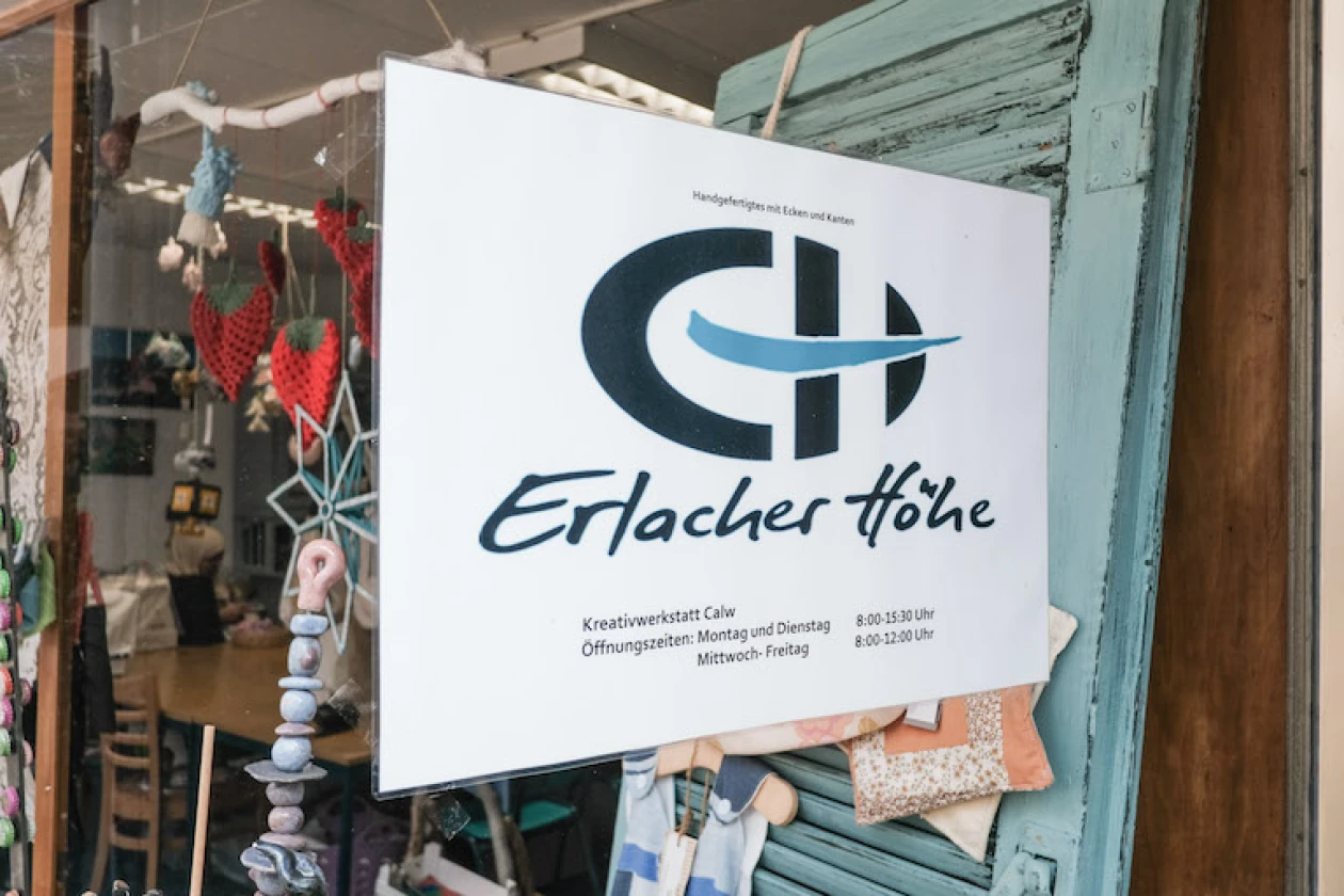 Ein Schild mit dem Logo und Firmennamen "Erlacher H&ouml;he" h&auml;ngt an einem Schaufenster, durch das man einen Blick auf einen Innenraum mit verschiedenen Dekoartikeln werfen kann.