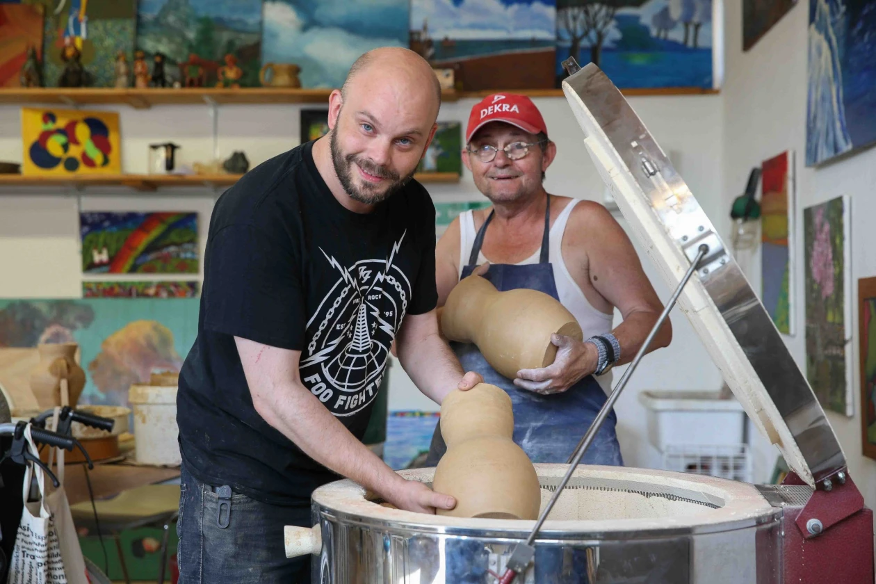 Zwei Personen stehen in einer Kreativwerkstatt mit zwei Tongef&auml;&szlig;en und blicken in die Kamera. Der eine tr&auml;gt ein schwarzes T-Shirt, der andere eine rote Kappe und eine blaue Sch&uuml;rze. Im Hintergrund sind bunte Kunstwerke und Gem&auml;lde zu sehen.