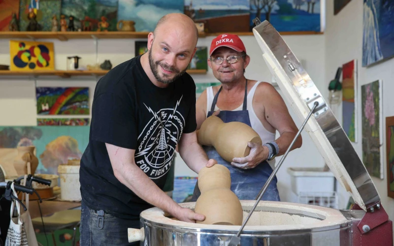 Zwei Personen stehen in einer Kreativwerkstatt mit zwei Tongef&auml;&szlig;en und blicken in die Kamera. Der eine tr&auml;gt ein schwarzes T-Shirt, der andere eine rote Kappe und eine blaue Sch&uuml;rze. Im Hintergrund sind bunte Kunstwerke und Gem&auml;lde zu sehen.