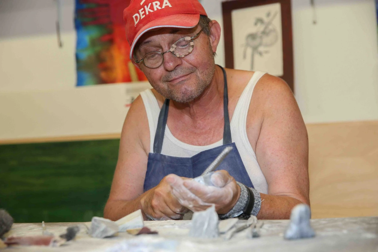 &Auml;lterer Mann mit Brille und roter Kappe arbeitet konzentriert an einer T&ouml;pferscheibe, tr&auml;gt eine blaue Sch&uuml;rze und formt mit seinen H&auml;nden einen Ton-Gegenstand.
