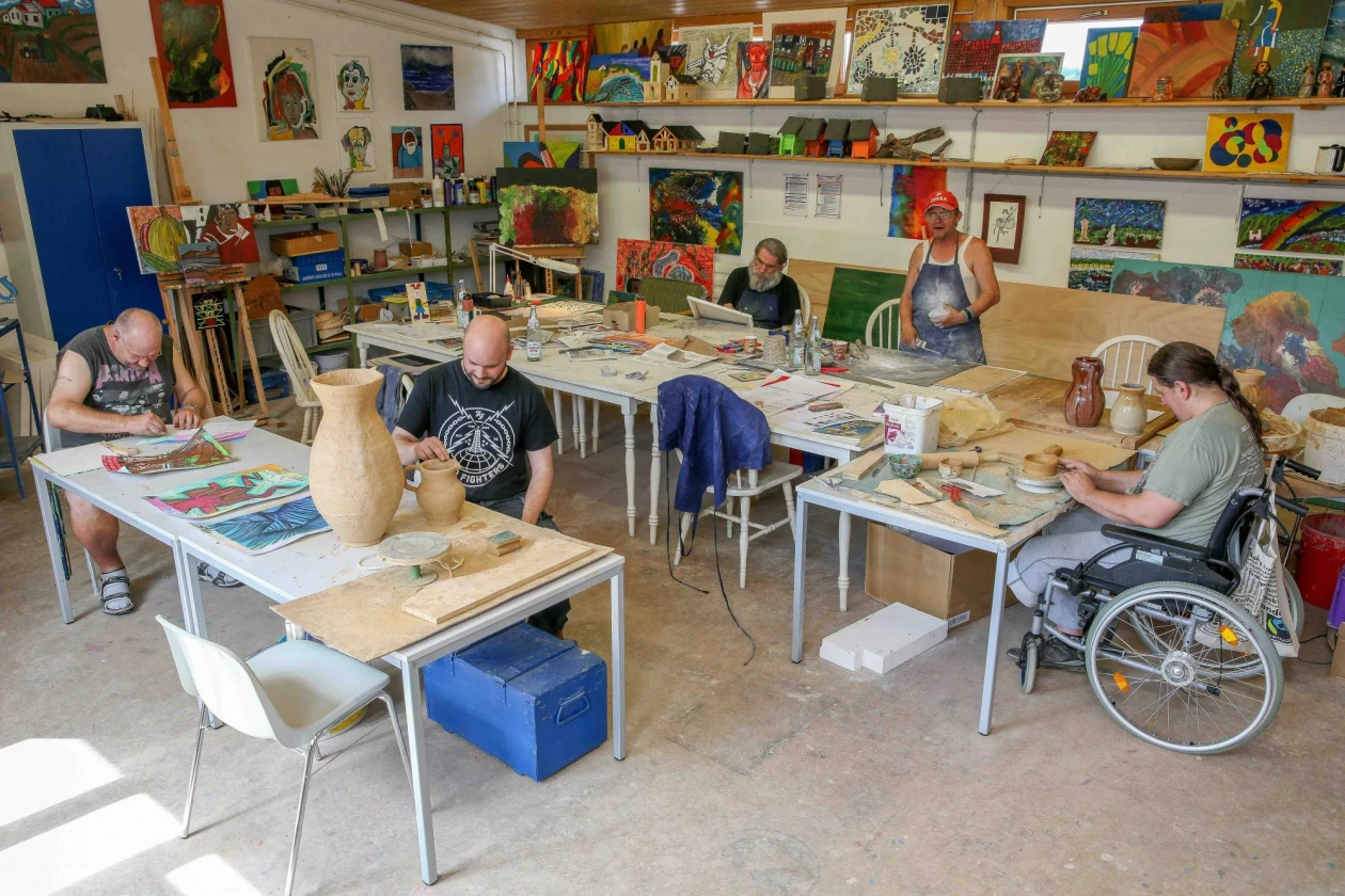 In einer hellen, farbenfrohen Kreativwerkstatt arbeiten f&uuml;nf Personen an Tischen an kreativen Projekten. Eine Person sitzt im Rollstuhl.