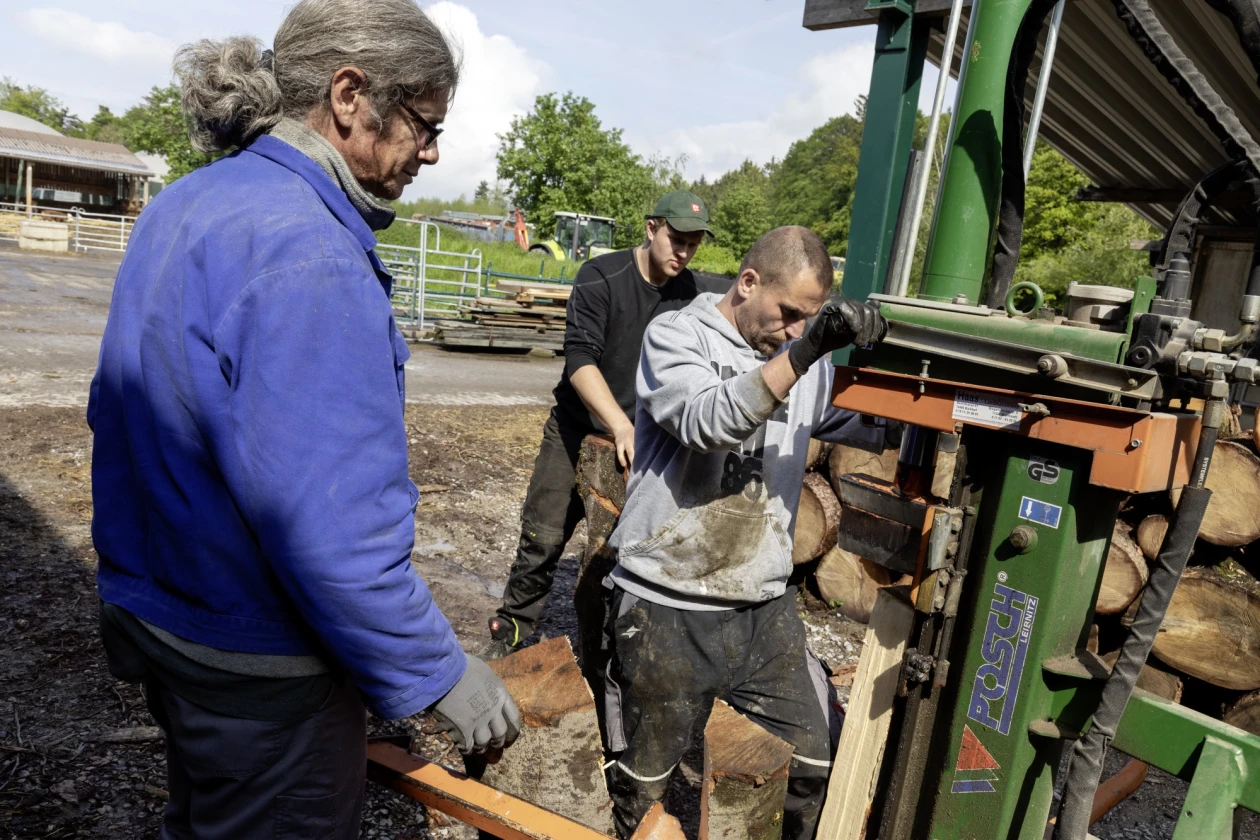 Drei M&auml;nner arbeiten an einer Holzbearbeitungsmaschine. Die Szene spielt sich auf einem Au&szlig;engel&auml;nde mit B&auml;umen und Z&auml;unen im Hintergrund ab.