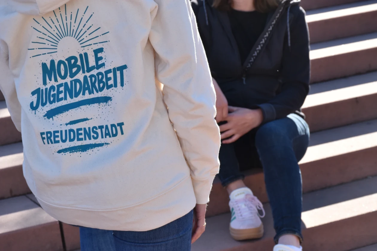 Im Vordergrund steht eine Streetworkerin mit dem R&uuml;cken zu und auf ihrem Pullover steht "Mobile Jugendarbeit Freudenstadt", sie spricht mit einem M&auml;dchen im Hintergrund welches auf der Treppe sitzt.