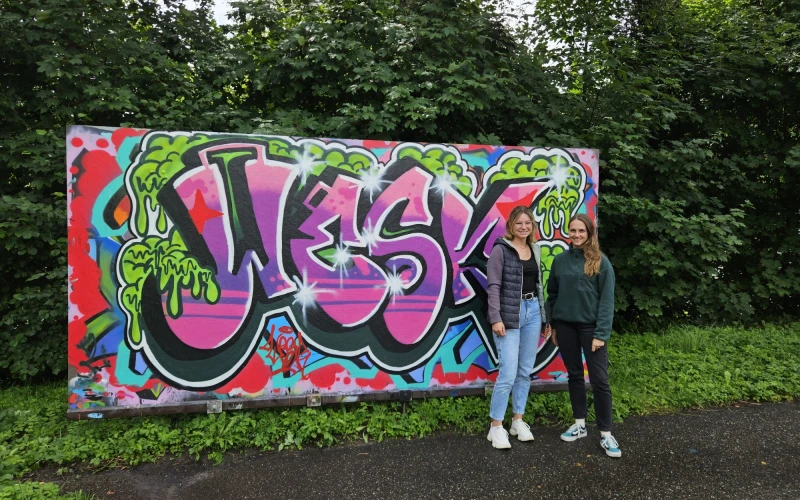 Zwei Personen stehen vor einer bunten Graffiti-Wand mit gro&szlig;fl&auml;chigen pinken und gr&uuml;nen Buchstaben sowie farbigen Verzierungen, umgeben von gr&uuml;nen Hecken und B&auml;umen.