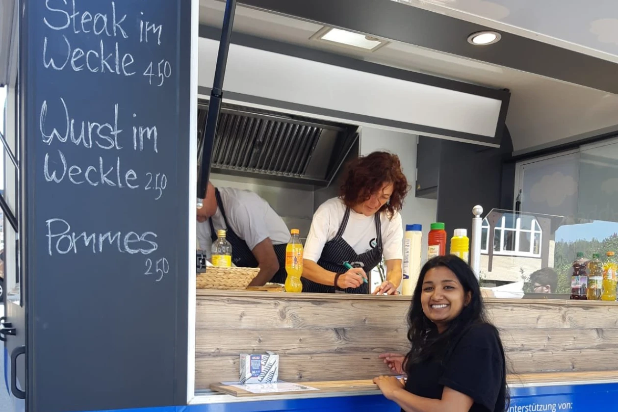Eine Frau l&auml;chelt vor dem Anh&auml;nger "Windr&auml;dle" der Erlacher H&ouml;he, w&auml;hrend hinter dem Tresen zwei weitere Personen zu sehen sind.