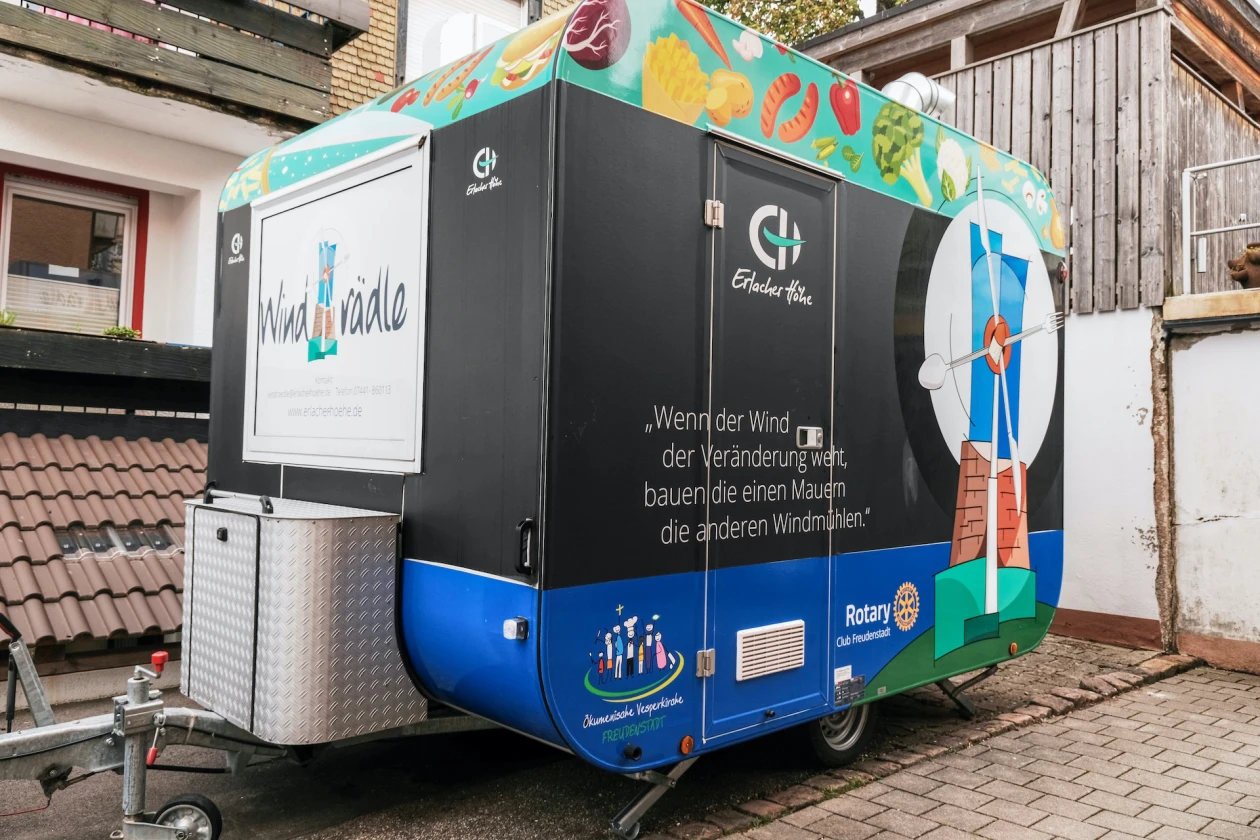 Ein mobiler Anh&auml;nger in Schwarz und Blau mit buntem Dach, beklebt mit der Aufschrift "Windr&auml;dle",  steht auf einem gepflasterten Hof.