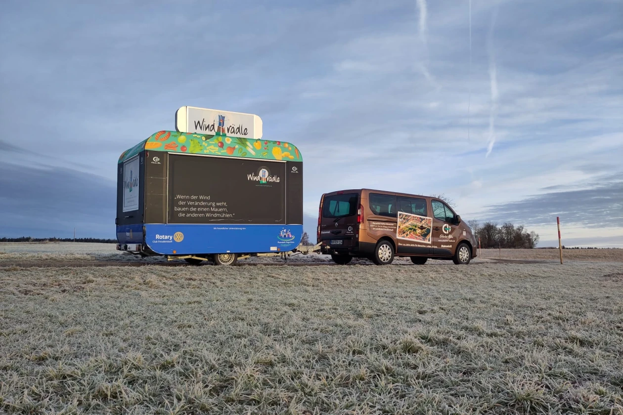 Unser braunes Firmenfahrzeug hat an der Anh&auml;ngerkupplung unseren mobilen Essenwagen. Wir wintern unseren Anh&auml;nger "Windr&auml;dle" ein. Im Hintergrund blauer Himmel und eine Wiese bedeckt mit Tau.