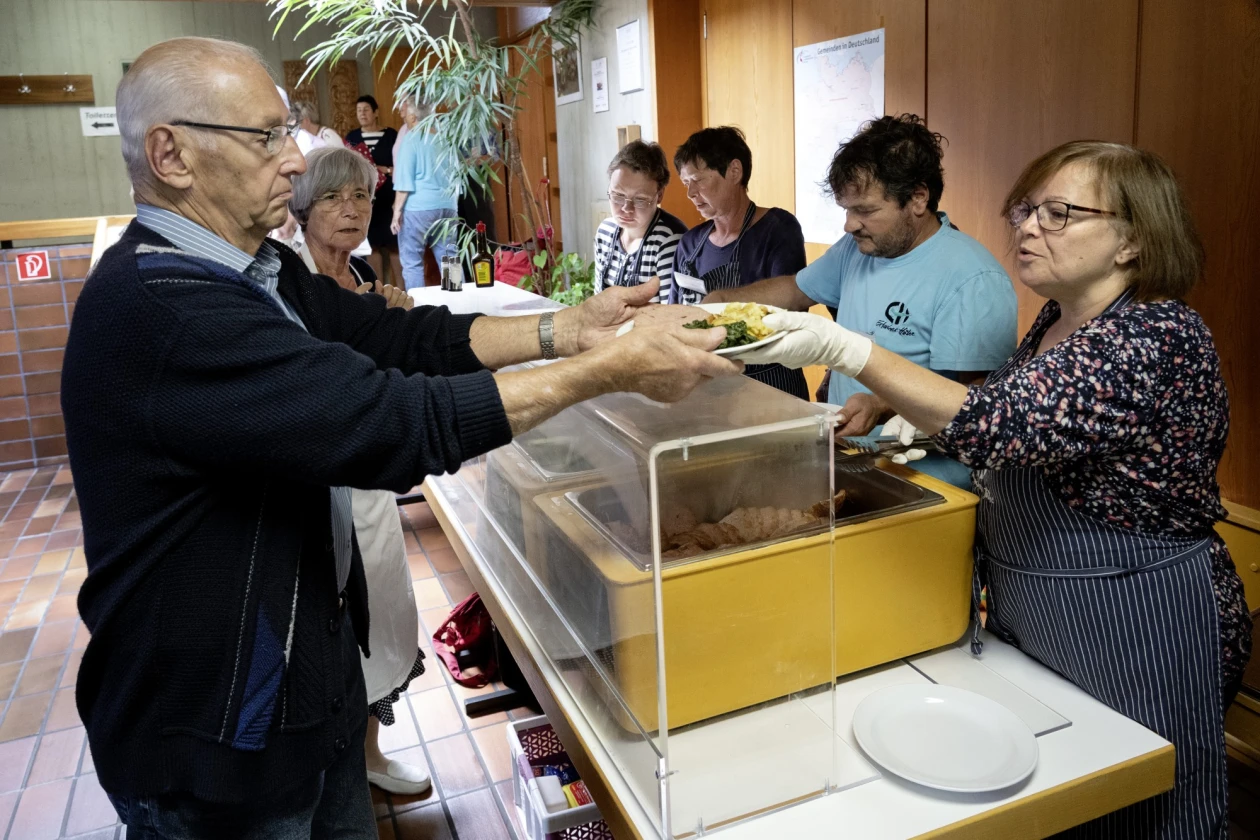 Bei einer Essensausgabe nimmt ein &auml;lterer Mann einen Teller mit Fleisch und Kartoffeln von einer Frau entgegen. Mehrere Personen stehen im Hintergrund und beobachten die Szene.