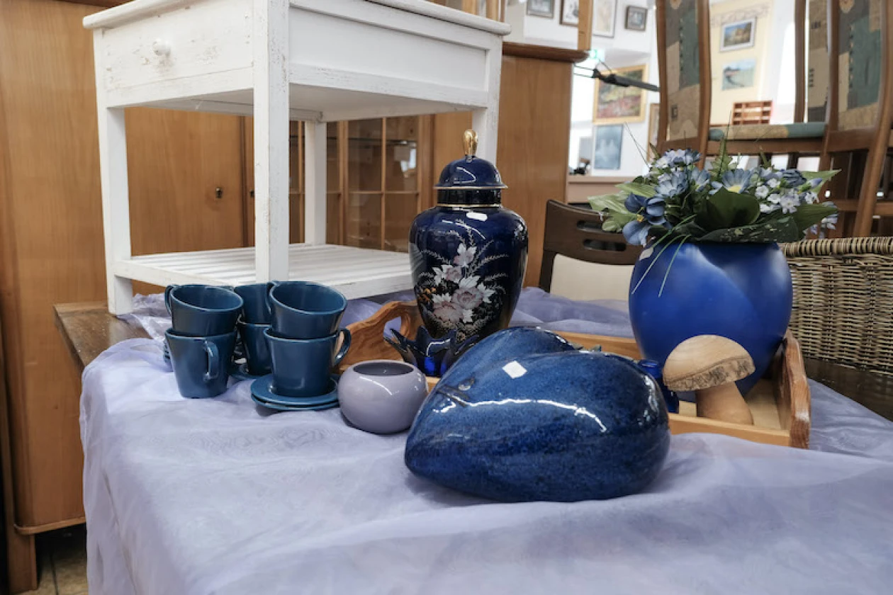 Auf einem Tisch stehen verschiedene blaue Keramikobjekte &ndash; darunter Tassen, Vasen, ein Herz sowie ein &Uuml;bertopf mit Kunstblumen. Im Hintergrund sind M&ouml;belst&uuml;cke und gerahmte Bilder an der Wand zu erkennen.