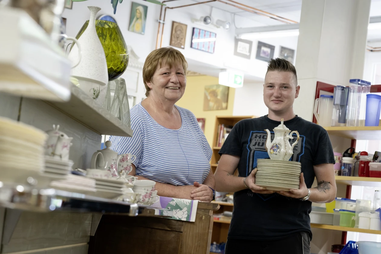 Zwei Personen blicken direkt in die Kamera: eine Frau in einem gestreiften Shirt lehnt l&auml;ssig an einem Schrank, w&auml;hrend ein junger Mann im schwarzen T-Shirt mehrere Teller und eine Teekanne in den H&auml;nden h&auml;lt. Im Hintergrund sind Haushaltswaren sowie Bilder an der Wand zu sehen.