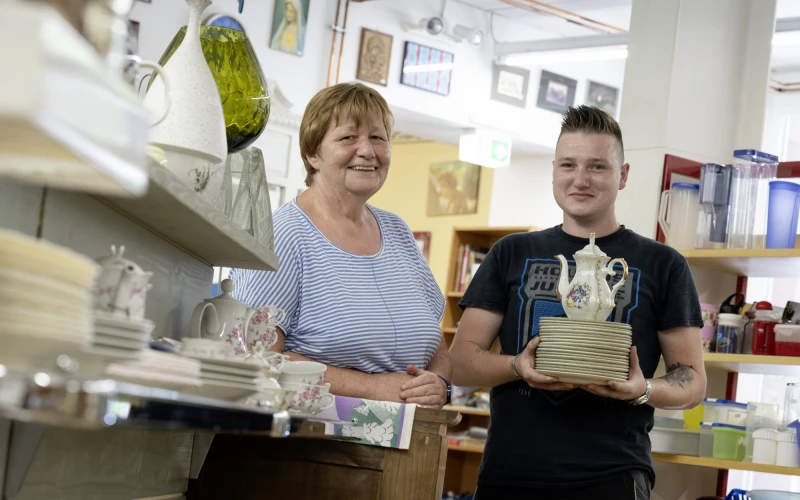 Zwei Personen blicken direkt in die Kamera: eine Frau in einem gestreiften Shirt lehnt l&auml;ssig an einem Schrank, w&auml;hrend ein junger Mann im schwarzen T-Shirt mehrere Teller und eine Teekanne in den H&auml;nden h&auml;lt. Im Hintergrund sind Haushaltswaren sowie Bilder an der Wand zu sehen.