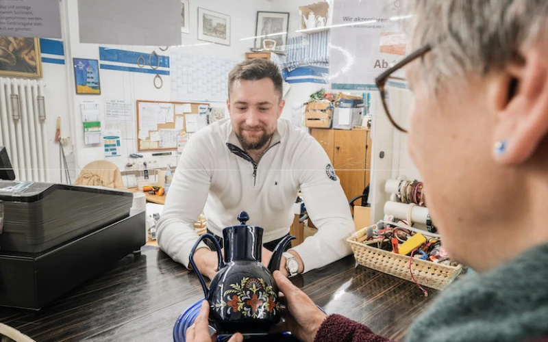 Ein Mann in einem wei&szlig;en Pullover steht l&auml;chelnd hinter der Kasse und blickt auf die Teekanne, die eine Frau vor ihm in den H&auml;nden h&auml;lt. Im Hintergrund sind Pinnw&auml;nde und Bilder an der Wand zu sehen.