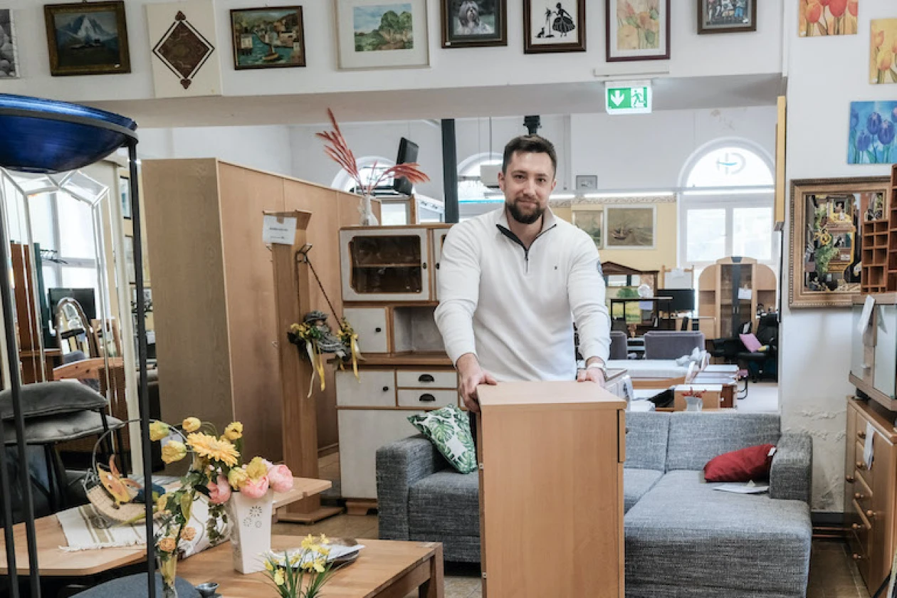 Ein Mann in wei&szlig;er Weste steht in einem Gebrauchtm&ouml;belladen und h&auml;lt mit beiden H&auml;nden einen Schrank fest. Im Raum befinden sich weitere M&ouml;belst&uuml;cke, Blumen auf einem Tisch sowie Bilder an den W&auml;nden.