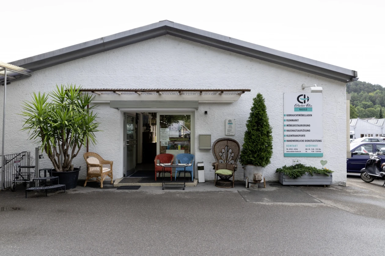 Wei&szlig;e Geb&auml;udefront mit Eingangst&uuml;r, davor stehen verschiedene Korbst&uuml;hle und eine Palme, rechts ein Firmenschild der Erlacher H&ouml;he. Ein Parkplatz ist im Hintergrund sichtbar.