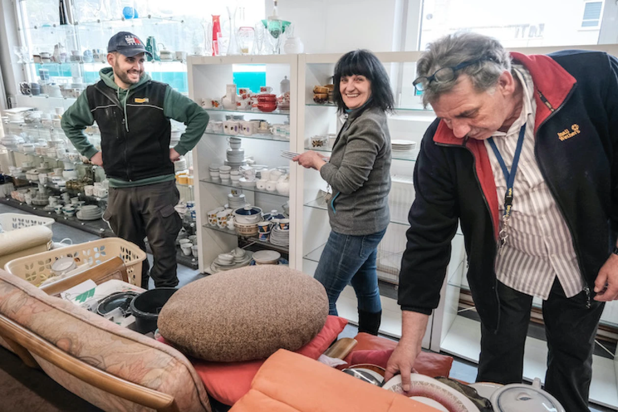 Drei Personen stehen in einem Sozialkaufhaus vor Regalen mit Geschirr. Die Personen scheinen bei der Arbeit oder Inventur zu sein.